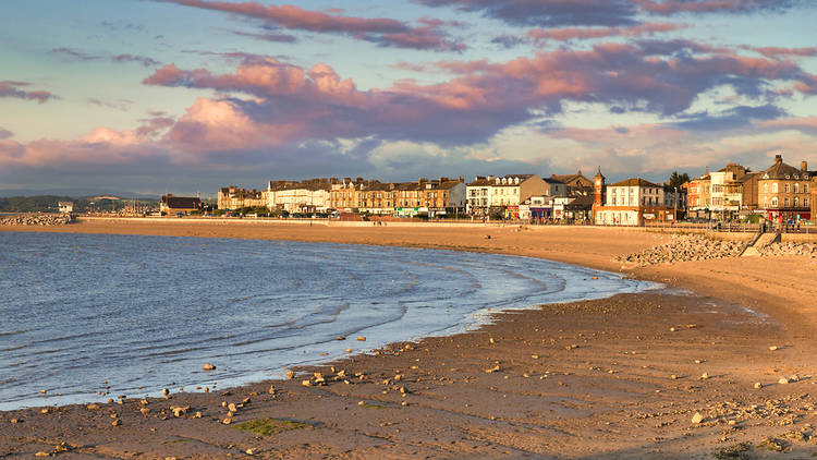 Golden hour at sunset on Morecambe Bay. A row of houses sit behind the curved yellow sand beach and purple clouds hang in the sky overhead.