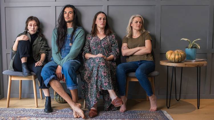 Four women and a man with long black hair sit on chairs in a row