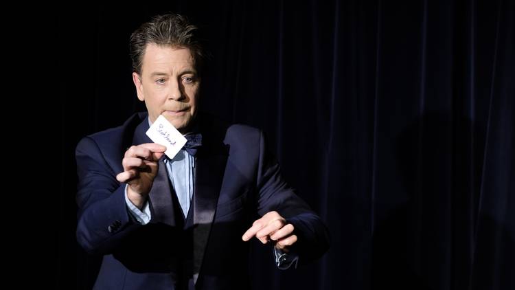 Magician Nick Nickolas wearing a suit and holding a card up