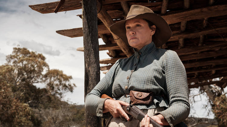 Still from The Drover’s Wife The Legend of Molly Johnson featuring Leh Purcell in cowboy hat and checked shirt holding a shotgun in a stable surrounded by gum trees