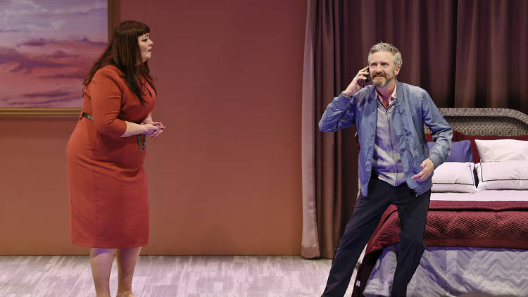 A man wearing a blue shirt and pants comically answers the phone while a woman wearing a red dress looks on concerned