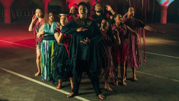 A group of people with brown skin and adorned with facial tattoos common to Maori and Pasifika cultures wear various flowing costumes and strike poses in a huddle facing forward