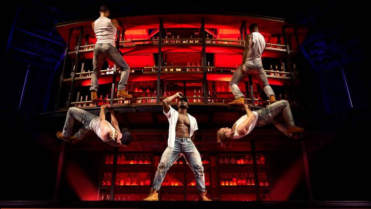 Five men clamber and dance on a red back-lit bar. The men are very athletic looking and are wearing white singlets with ripped jeans
