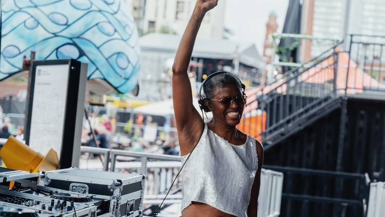 DJ Paulette is a black woman with cropped hair. She is wearing headphones and stands on stage before a mixing deck, smiling, her arm raised