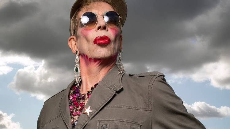 David Hoyle wearing dark glasses and red lipstick against a stormy clouded sky