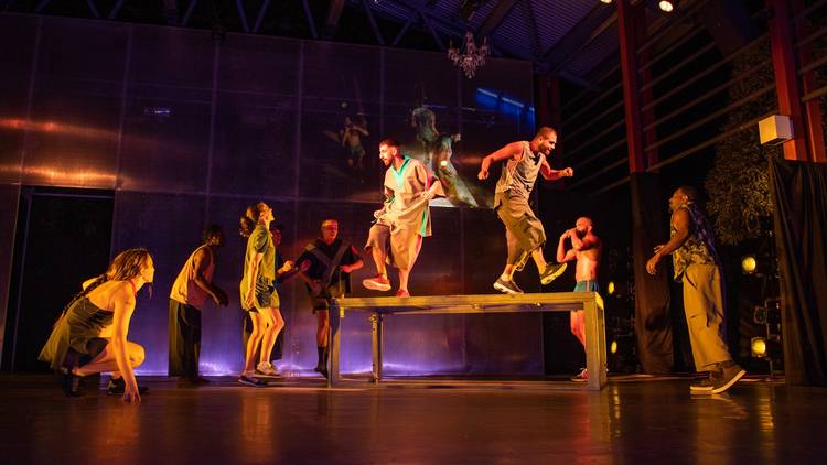 A group of 8 dancers performing around a table. Two of them, men wearing baggy street clothes, are dancing on the table