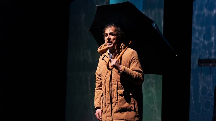 A middle aged man wearing a brown parka holds a black, cane-handled umbrella over his head