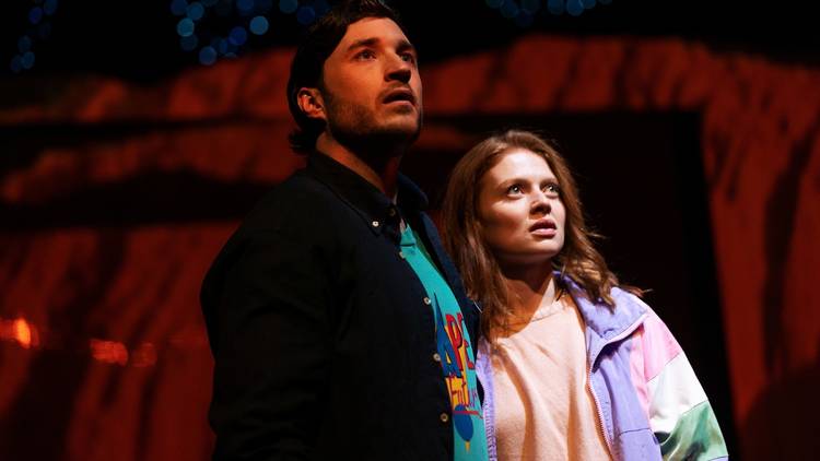 A man wearing a dark shirt and a woman wearing a light pink shirt and jacket look up in astonishment at something unseen