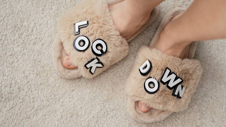 Close up of a pair of feet wearing fluffy beige slippers. The word "lockdown" is spelled out with plastic letters on the slippers.