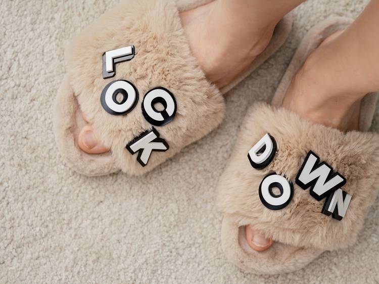 Close up of a pair of feet wearing fluffy beige slippers. The word "lockdown" is spelled out with plastic letters on the slippers.