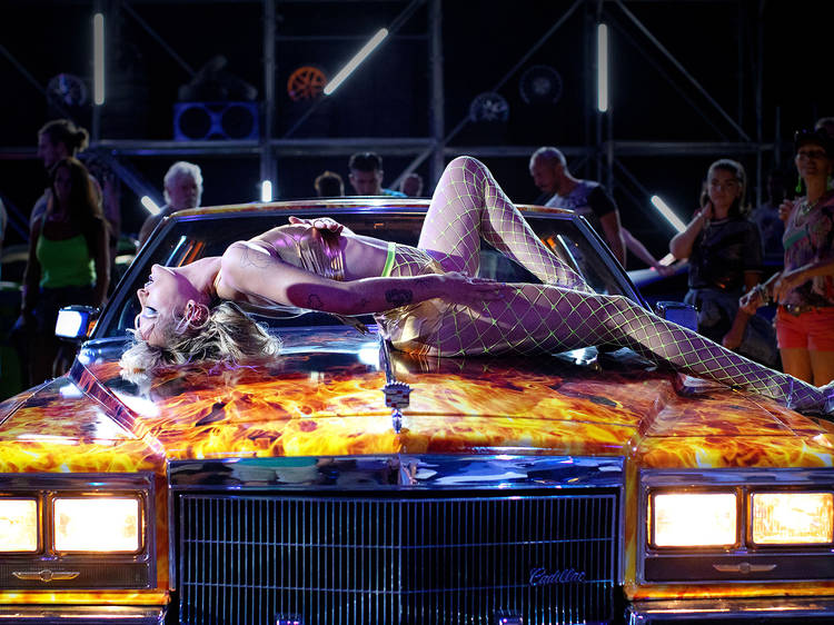 A blone woman in a corset and suspenders lies on the bonnet of a racer car with fire painted on it