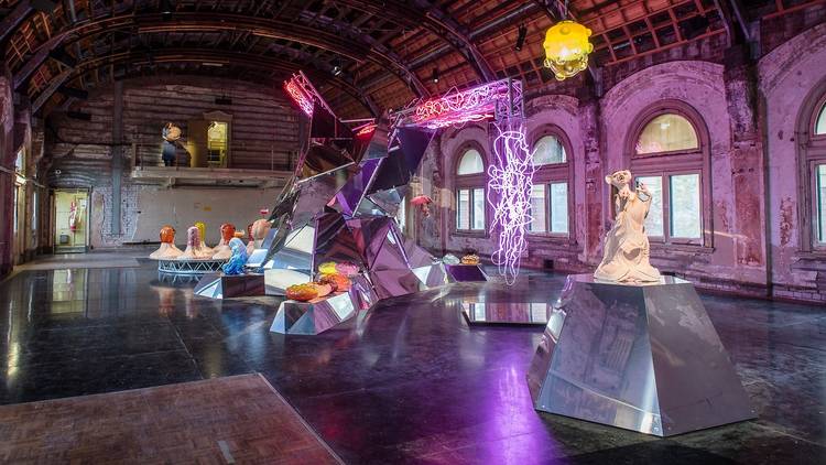 A large, decrepit  ballroom filled with shiny metal plinths and sculptures. There is a large pink neon light tangle dangling in the mid ground, while a large skinny rat sculpture sits on a plinth.