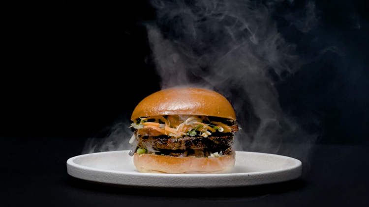 A brioche bun with pickled carrots and mushroom patty on a white plate surrounded by smoke whisps