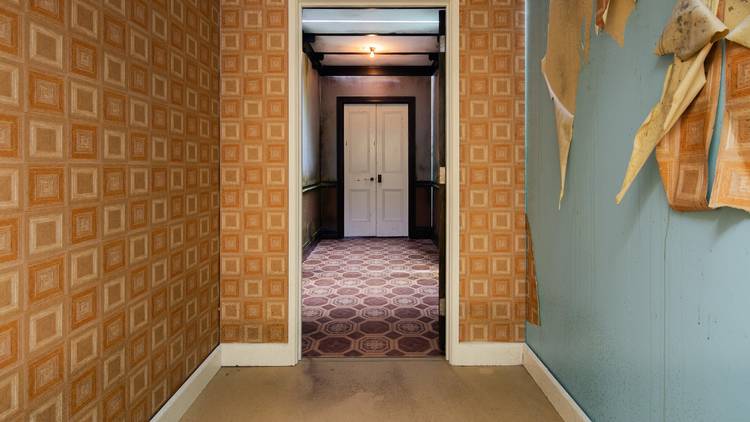 A hallway looking towards a door; some of the wallpaper is peeling