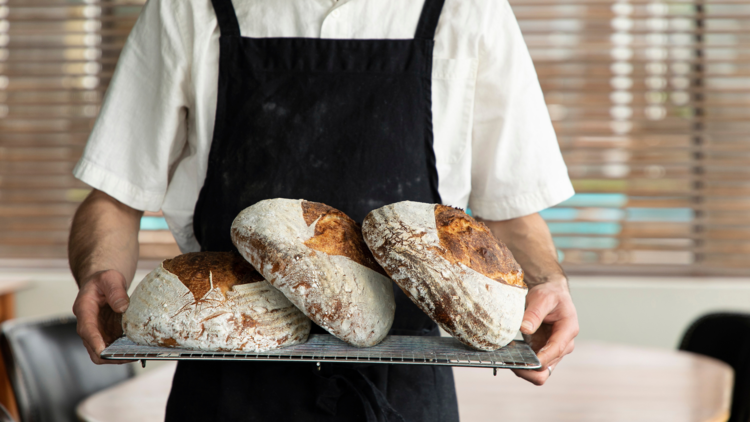 A baker with a black apron holds a cooling rack with three loaves of bread