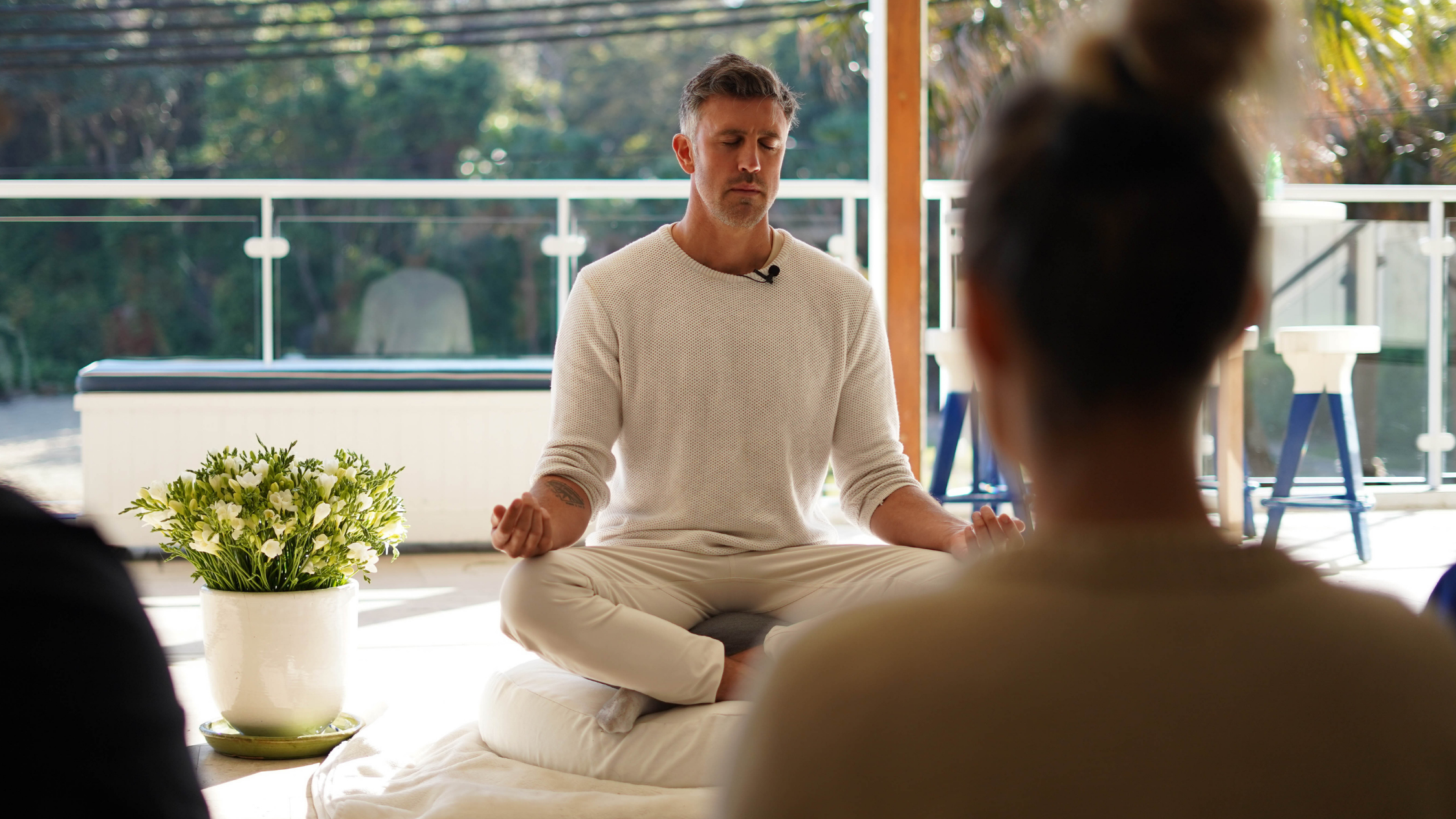 Luke McGregor instructs a meditation class. He wears a white jumper and pants and and is sitting cross-legged with his eyes clothes.