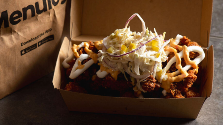 A cardboard take away box with fried chicken, creamy light slaw and diced pickles. Menulog paper bag in background.