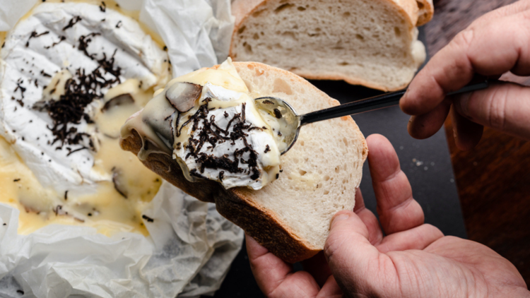 Hands hold a piece of sourdough with truffle brie being spooned on