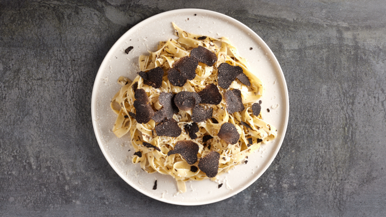 White plate with pile of pasta in centre with rounds of shaved truffle over the top. Slate table
