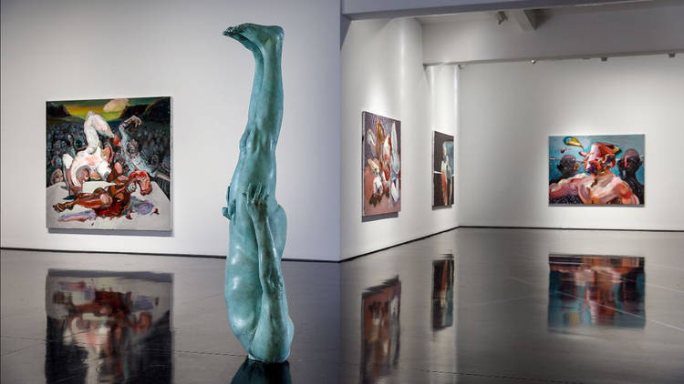 A gallery filled with large abstract paintings of men fighting. A upside down bronze sculpture of a naked man is in the foreground