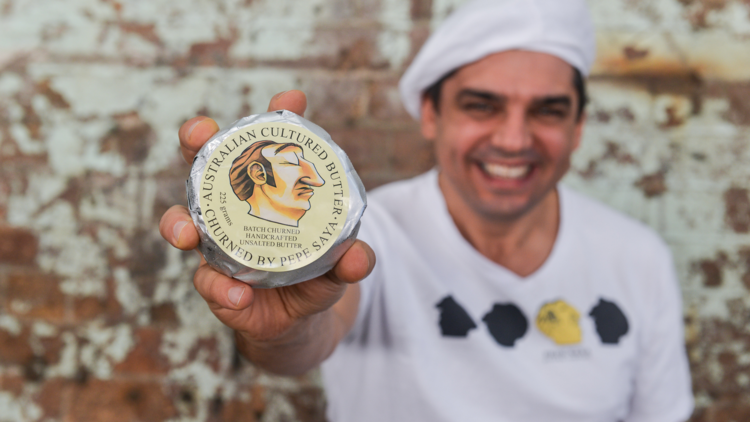 In focus a pat of Pepe Saya butter being held but a man against a rustic wall