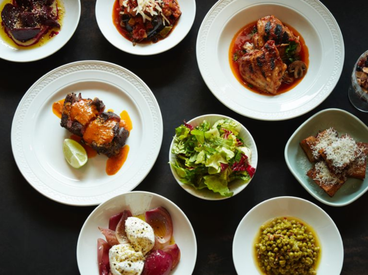 A slate table with a flat lay of multiple round plates, salad, braised meats, mozzarella and peas