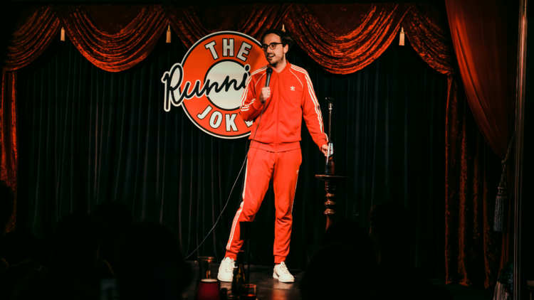 Comedian Daniel Muggleton wears a red tracksuit and stand on stage with a microphone.