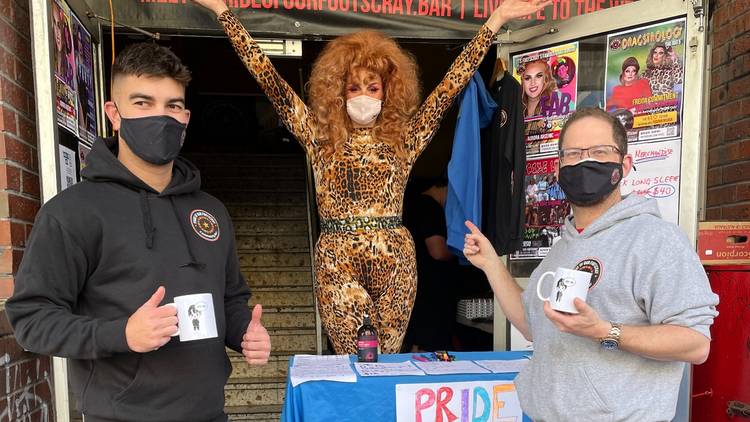 Two people wearing masks smile after getting drinks from a drag queen