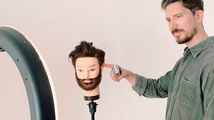 Barber Nic Singer demonstrates how to give a home haircut, he is holding an electric razor and pointing at a dummy head with beard.