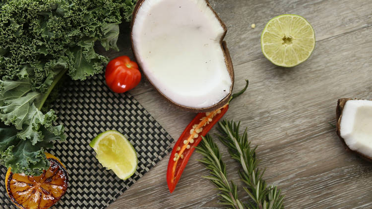 Half a coconut is placed on a wooden surface next to limes, herbs and a chilli