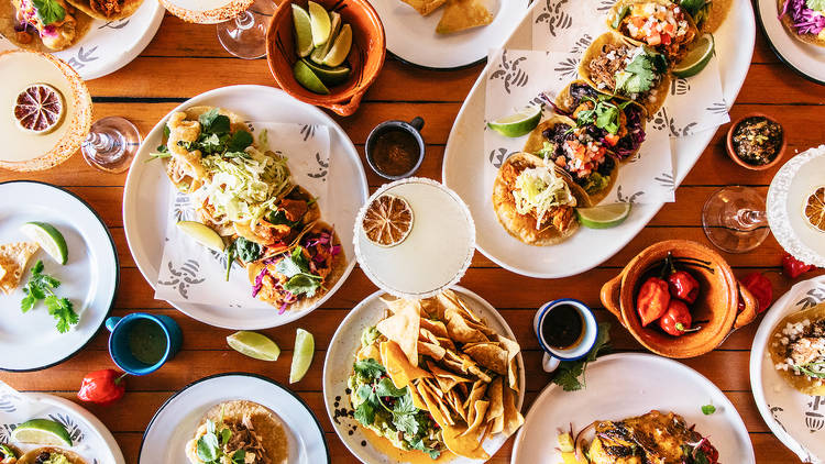 Multiple trays of tacos on a wooden table with a margarita and bowls of chips