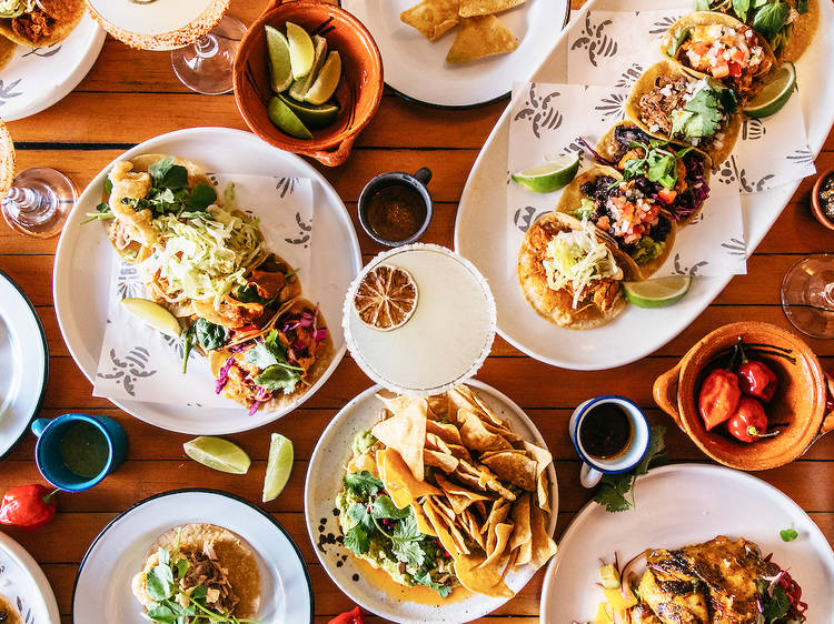 Multiple trays of tacos on a wooden table with a margarita and bowls of chips