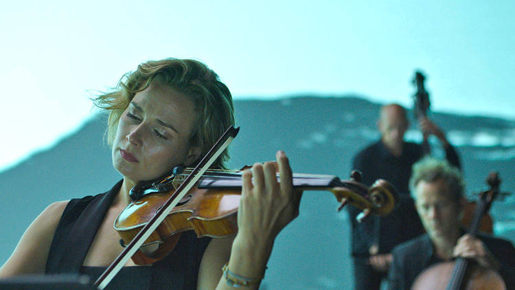 A woman in the Australia Chamber Orchestra playing the violin against a bright blue video backdrop of waves