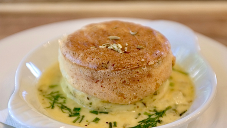 A crab souffle on a white plate with a butter sauce and herbs