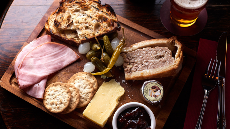 A ploughmans board of ham, cheese, preserves and pork pie on a dark wooden table with a pint of beer