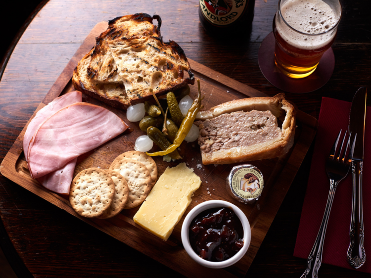 A ploughmans board of ham, cheese, preserves and pork pie on a dark wooden table with a pint of beer