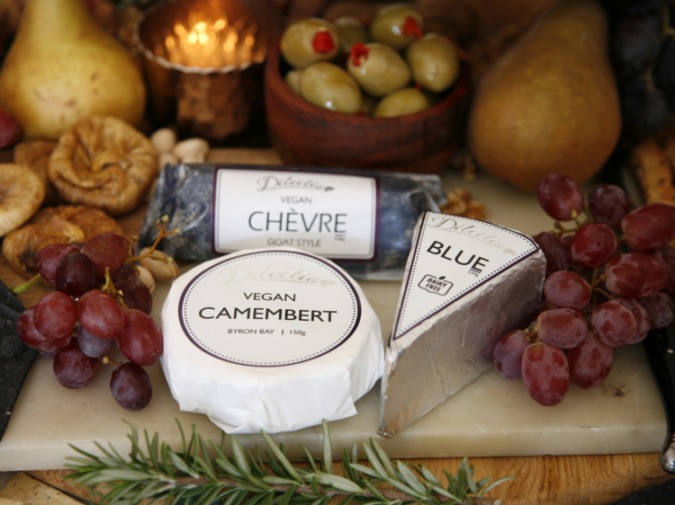 Three packaged vegan cheeses are displayed on a board with grapes and figs.