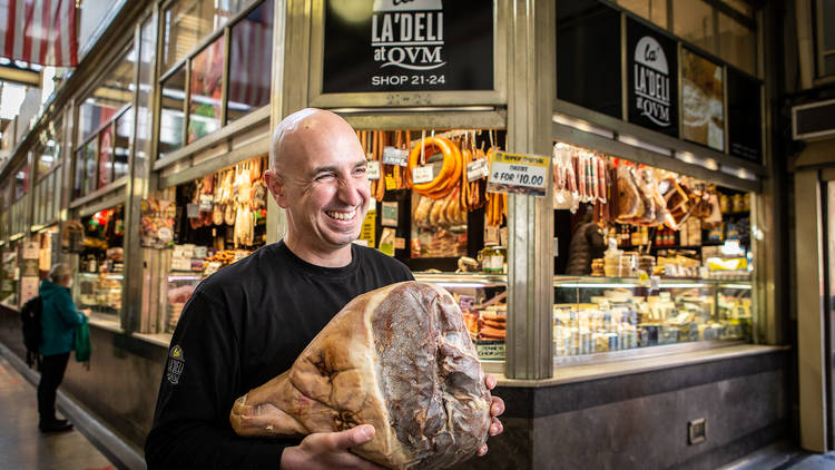 A man holding a cut of meat in front of a deli. 