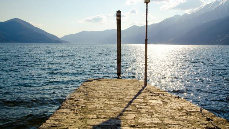 A small jetty leads into a lake with a lamp post at the end and views of mountains and sky in the distance.