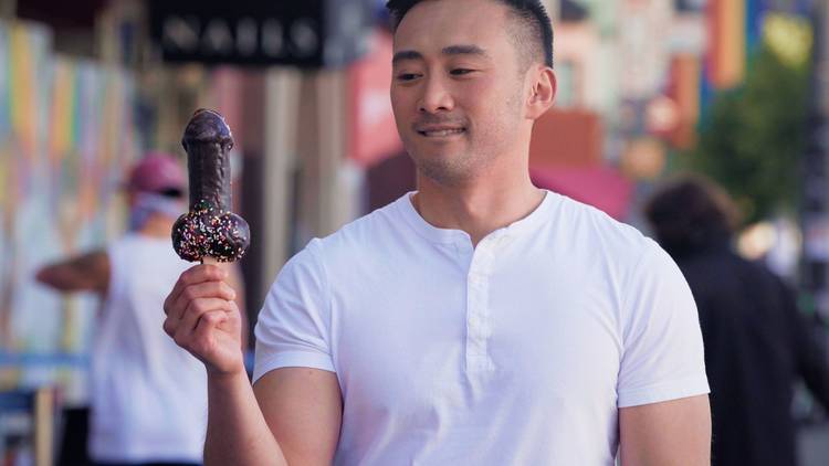 A cute man in a white t-shirt looks at a chocolate penis lolly