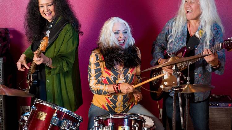 Filipina American rock band Fanny in cool clothes playing instruments against a pink backdrop 