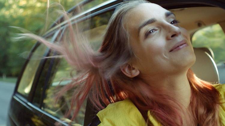 A woman in a yellow shirt with died pink hair leans out a car window in the film Lola