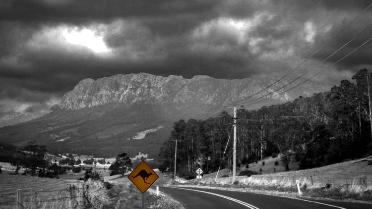 A black and white image of a country road with a towering mountain range and a kangaroo crossing road sign in yellow