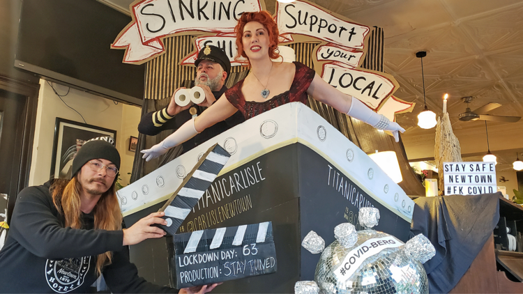 Two actors and a person holding a clapperboard are filming a tribute to Titanic in a bar with a replica ship.