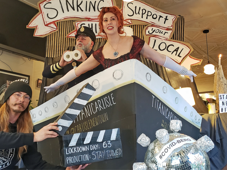 Two actors and a person holding a clapperboard are filming a tribute to Titanic in a bar with a replica ship.