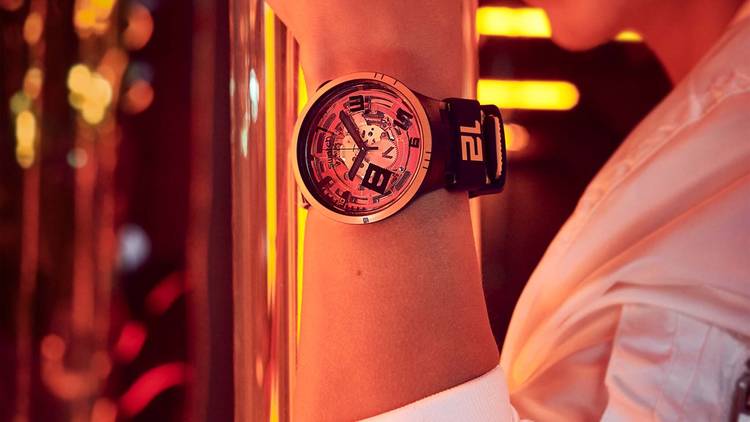 The bright orange Oops! Swatch watch shown on an arm with the person's face seen smiling in the back of the frame. Orange neon lights and reflective surfaces can be seen in the background.