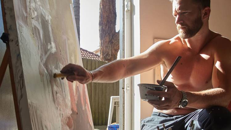 Actor Tim Draxl sitting at an easel painting, bare chested
