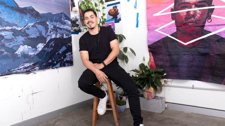 Artist Abdul Abdullah in black t-shirt and jeans sitting on a stool in front of hist artwork