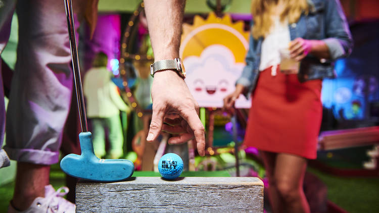 A blue golf ball is placed on a wooden block, with a blue golf club next to it, ready to strike