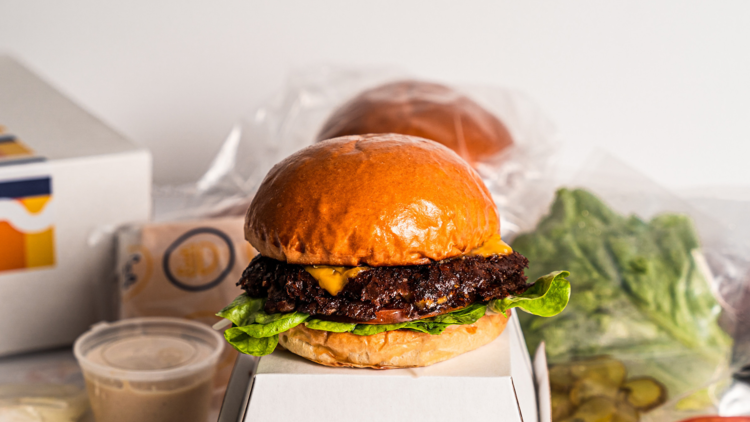 A burger sits atop a white box, vac packed ingredients in background